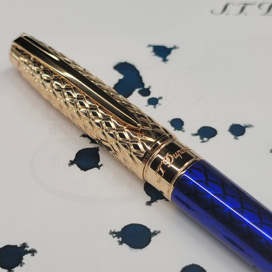 Luxury rollerball pen with intricate rose gold guilloche cap and deep blue textured barrel on ink-splattered paper