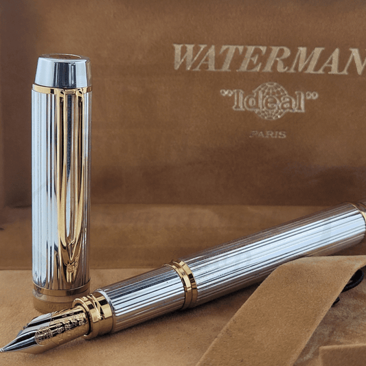 Waterman Le Man 100 Sterling Silver fountain pen with vertical line pattern, gold-tone trim, 18K medium nib, shown disassembled with cap and barrel against vintage Waterman logo