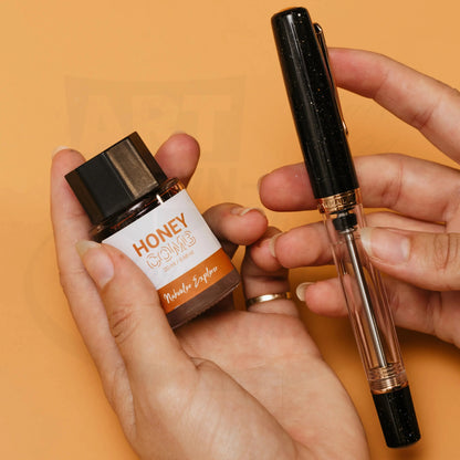 Nahvalur Original Plus Lovina Graphite fountain pen in resin with rose gold trim, displayed with Honey ink bottle on orange background