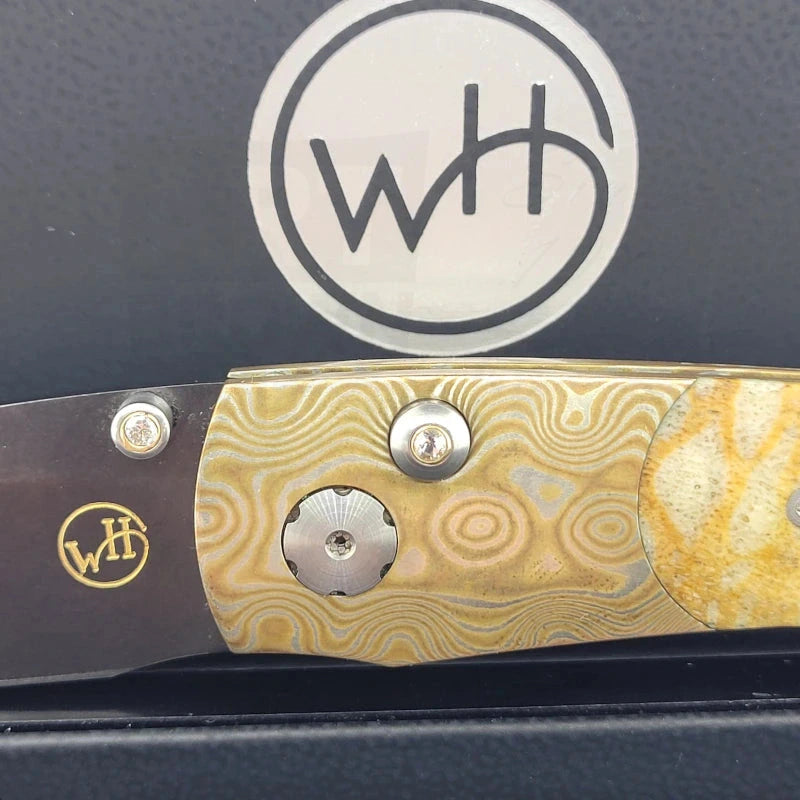 Detailed view of the William Henry B7 knife's Mokume-Gane frame and lock button adorned with white topaz, set against the dark interior of the box